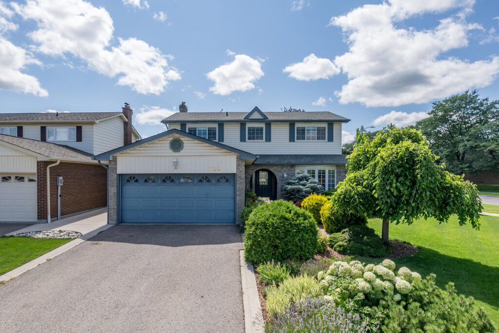 Two storey house for sale in Milton with double car garage. A blue sky with puffy white clouds above, and green grass with small trees in front.